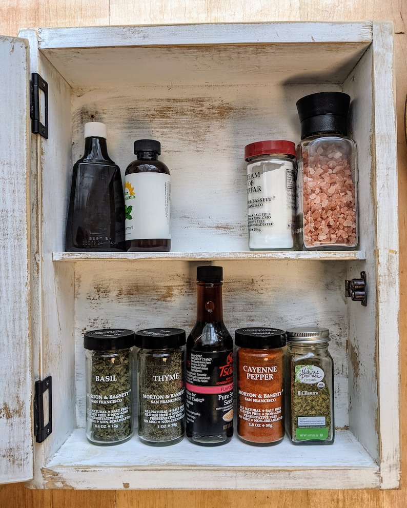 May include: A white, distressed wooden spice rack with two shelves. The top shelf holds two bottles and two jars. The bottom shelf contains four jars of spices and a bottle of soy sauce. The rack is open, revealing the contents.