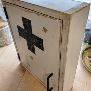 May include: A distressed, off-white wooden cabinet with a black cross emblem on the front. The cabinet has a hinged door with a black handle and is designed to be mounted on a wall. The cabinet's finish has a rustic, aged appearance.