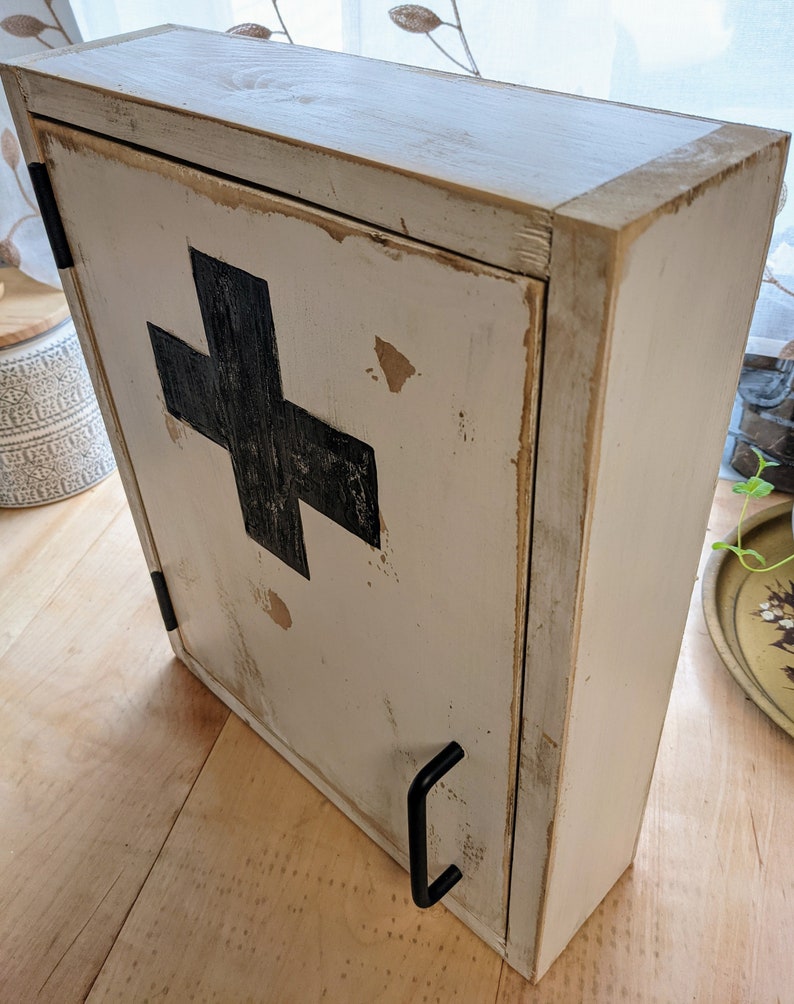May include: A distressed, off-white wooden cabinet with a black cross emblem on the front. The cabinet has a hinged door with a black handle and is designed to be mounted on a wall. The cabinet's finish has a rustic, aged appearance.
