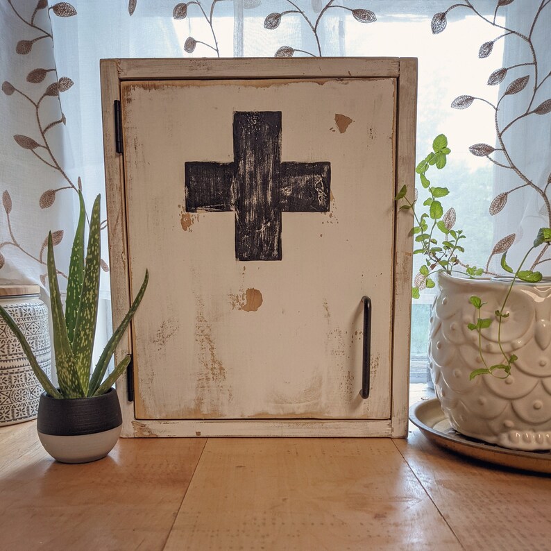 May include: A distressed white wooden cabinet with a black cross emblem on the front. The cabinet has a black handle and is placed on a wooden surface. A potted aloe plant and a decorative owl planter are visible.