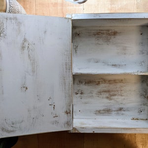 May include: A small, white, distressed wooden cabinet with a single door open to reveal two shelves. The cabinet has a rustic, aged appearance with visible wood grain and brown undertones peeking through the white paint. The door is attached with a black hinge.