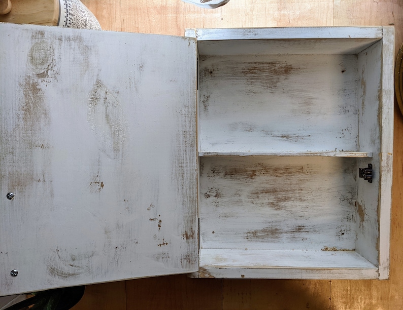May include: A small, white, distressed wooden cabinet with a single door open to reveal two shelves. The cabinet has a rustic, aged appearance with visible wood grain and brown undertones peeking through the white paint. The door is attached with a black hinge.