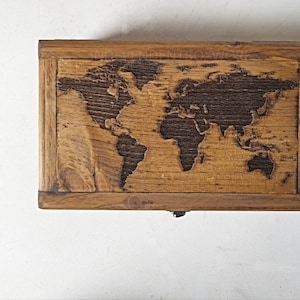May include: A rectangular wooden box with a world map carved into the lid. The box is made of light brown wood with darker brown accents. The map shows the continents and oceans in a detailed relief.