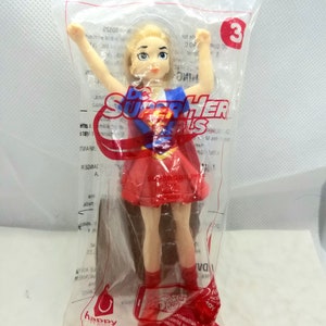 Puede incluir: Una figura de juguete de plástico roja y azul de una superheroína con cabello rubio, que lleva una falda roja y botas rojas. La figura tiene los brazos en alto. El embalaje es de plástico transparente con el número 3 impreso en él. El texto "DC Super Hero Girls" está impreso en el embalaje.