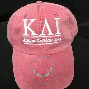 May include: A pink, distressed, cotton baseball cap with white embroidery that reads "ΚΛΙ kappa lambda iota". The cap has a white stitching detail around the brim and a small embroidered logo on the back.