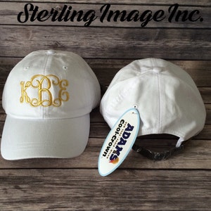May include: Two white baseball caps with a gold embroidered monogram on one cap. The cap on the right has a white tag with the text "ADAMS Coolcrown" in blue and red.