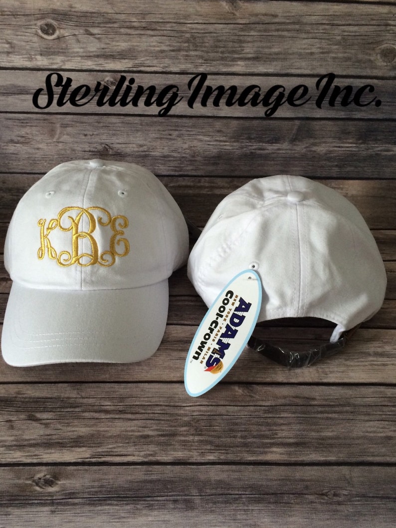 May include: Two white baseball caps with a gold embroidered monogram on one cap. The cap on the right has a white tag with the text "ADAMS Coolcrown" in blue and red.