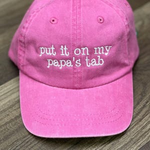 May include: A pink baseball cap with the embroidered text "put it on my papa's tab" in white. The cap has a curved brim and a fabric strap closure. The hat is made of a soft, washed cotton material.