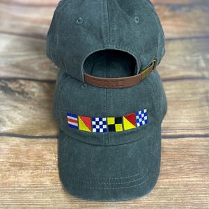 May include: A dark charcoal gray baseball cap featuring an embroidered row of six colorful nautical signal flags on the front panel. The cap has a curved brim, stitched eyelets, and an adjustable brown leather strap with a metal buckle at the back.