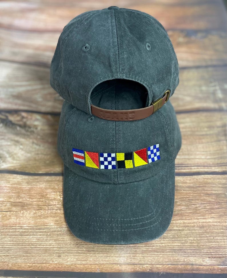 May include: A dark charcoal gray baseball cap featuring an embroidered row of six colorful nautical signal flags on the front panel. The cap has a curved brim, stitched eyelets, and an adjustable brown leather strap with a metal buckle at the back.