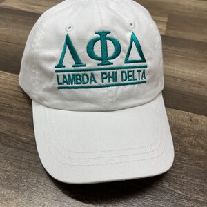 May include: A white baseball cap with a curved brim. The cap features teal embroidery of Greek letters and the words "LAMBDA PHI DELTA" across the front. The cap has a button on top and adjustable strap.