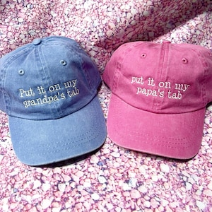 May include: A blue baseball cap with the white embroidered text "Put it on my grandpa's tab" and a pink baseball cap with the white embroidered text "put it on my papa's tab". Both caps feature a curved brim, a button top, and eyelet vents.