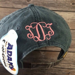 May include: A dark blue denim baseball cap with a pink embroidered monogram. The cap has a brown leather strap and a white label with the text "Adams Cool-Crown" in blue and red.