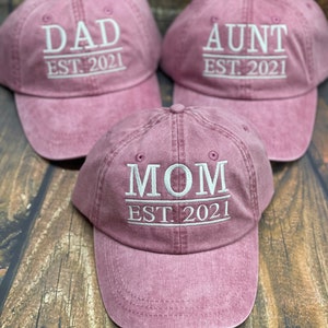 May include: Three pink baseball caps with white embroidered text. The caps read "DAD EST. 2021", "MOM EST. 2021", and "AUNT EST. 2021".