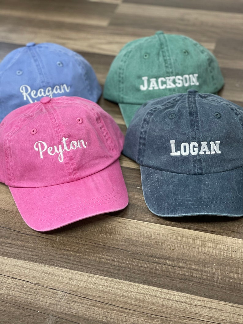 May include: Four personalized baseball caps in different colors: blue, pink, green, and dark blue. The caps have the names "Reagan", "Peyton", "Jackson", and "Logan" embroidered on them in white.