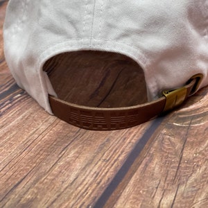 May include: White baseball cap with a brown leather strap that says "PALM" in raised lettering.