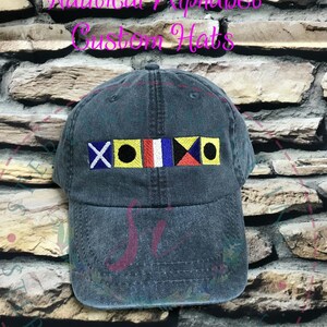 May include: A dark blue denim baseball cap with a row of colorful nautical signal flags embroidered on the front. The flags are red, yellow, blue, white, and black. The text "Nautical Alphabet Custom Hats" is written in pink above the flags.