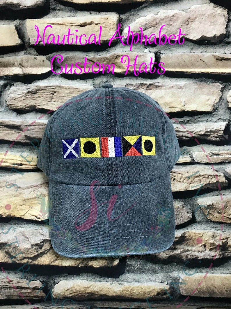 May include: A dark blue denim baseball cap with a row of colorful nautical signal flags embroidered on the front. The flags are red, yellow, blue, white, and black. The text "Nautical Alphabet Custom Hats" is written in pink above the flags.