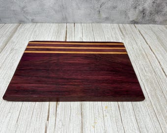 May include: A rectangular wooden cutting board with a deep purple hue. Two thin, parallel, light-brown stripes are inlaid near the top edge. The board is set on a light-colored, wooden surface.