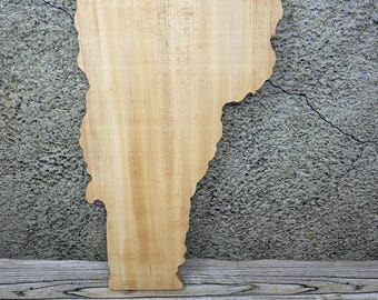 May include: A wooden cutout in the shape of the state of Vermont. The wood has a light, natural color with visible grain patterns. The cutout is displayed against a textured gray wall and a wooden surface.