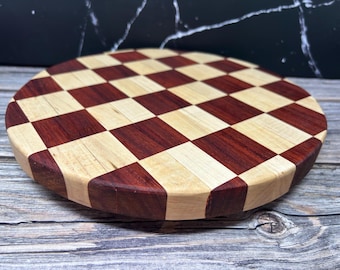 May include: Round wooden cutting board with a checkerboard pattern. The alternating squares are light and dark wood tones, creating a visually appealing design. The board is likely used for food preparation.
