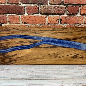 May include: A rectangular wooden serving board with a river of blue resin flowing through the center. The wood has a natural grain pattern, and the resin has a glossy finish. The board is displayed against a brick wall.