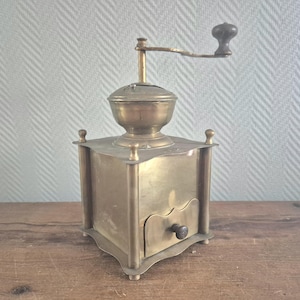 Puede incluir: Antiguo molinillo de café de latón con manivela de madera. El molinillo tiene una base cuadrada con un cajón y una parte superior decorativa. El latón presenta una pátina cálida y envejecida. Una herramienta de cocina clásica.