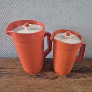 Può includere: Due brocche di plastica arancione con coperchi e manici bianchi. Le brocche sono di dimensioni diverse, la più grande a sinistra. I coperchi hanno un piccolo pomello bianco in cima. Sono su una superficie di legno.