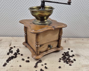 Könnte beinhalten: Eine antike Kaffeemühle aus Holz mit einem Messingbehälter und einer Handkurbel. Die Mühle hat eine Schublade zum Aufbewahren von Kaffeebohnen und steht auf einer Holzoberfläche mit verstreuten Kaffeebohnen.