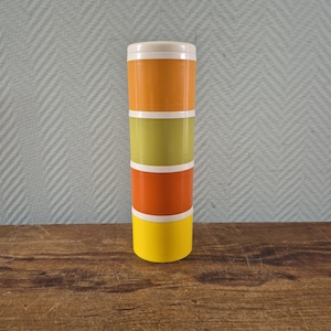 Puede incluir: Un recipiente de alimentos apilable de estilo retro, con tapa blanca. Consta de cuatro secciones en naranja, verde oliva, naranja y amarillo, separadas por bandas blancas. El recipiente es de plástico y está sobre una superficie de madera.
