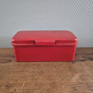 May include: A red metal storage box with a hinged lid. The box is rectangular and has a slightly rounded top.