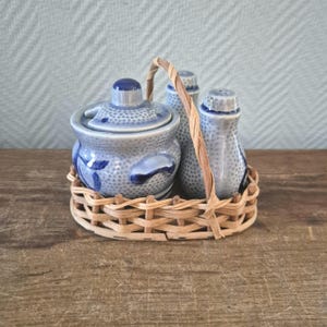 Op de afbeelding: Een blauw-wit keramisch kruidenset in een gevlochten mand. De set bevat een pot met deksel, een zoutstrooier en een peperstrooier. De keramische stukken hebben een stippenpatroon en bloemmotieven. De mand heeft een handvat.