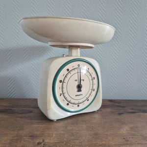 Op de afbeelding: Een witte vintage keukenweegschaal met een groene wijzerplaat. De wijzerplaat heeft zwarte markeringen en geeft tot 10 kg aan. De weegschaal is van Krups.