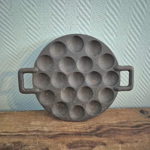 Op de afbeelding: Een ronde gietijzeren pan met meerdere cirkelvormige inkepingen, bedoeld voor het bakken van kleine pannenkoeken of gebakjes. De pan heeft twee rechthoekige handvatten en een donkere, matte afwerking. Hij staat op een houten oppervlak tegen een getextureerde, lichtblauwe achtergrond.