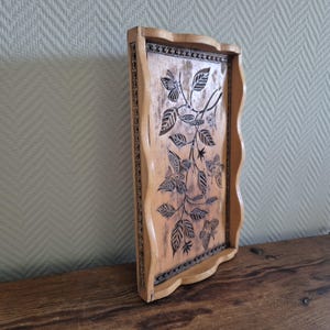 Op de afbeelding: Een houten dienblad met een gesneden bloemenmotief. Het dienblad is lichtbruin en heeft een golvende rand. Het ontwerp bevat bladeren, bloemen en vlinders.