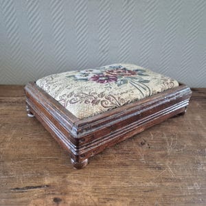 Peut inclure: Un repose-pieds rectangulaire en bois avec un coussin à motifs floraux. Le coussin présente un fond crème avec des motifs floraux bordeaux, verts et violets. Le repose-pieds est en bois brun foncé et possède de petits pieds arrondis.
