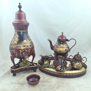Op de afbeelding: Een set van vijf handgeschilderde, rood- en gouden theepotten en een roomkan met ingewikkelde ontwerpen. De set bevat een grote theepot met een tuit en een handvat, een kleinere theepot met een tuit en een handvat, een roomkan met een handvat en twee kleine theekopjes. De set wordt gepresenteerd op een houten dienblad met een gouden rand.
