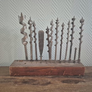 Op de afbeelding: Een verzameling antieke boren van verschillende maten en vormen, tentoongesteld in een houten houder. De boren zijn van metaal en vertonen tekenen van ouderdom en gebruik. De houten houder is roodbruin gekleurd.