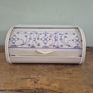 Op de afbeelding: Een vintage broodtrommel met een gebogen bovenkant en een witte handgreep. De trommel is lichtgrijs met een blauw bloemmotief over de voorkant. Het ontwerp omvat bloemen en ranken. De broodtrommel staat op een houten oppervlak.