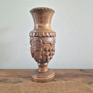 Puede incluir: Un jarrón de madera tallada con base de pedestal. El jarrón presenta un relieve detallado de uvas y hojas. El borde superior tiene un borde festoneado. La madera tiene un tono marrón cálido y el jarrón mide unos 20 cm de alto.
