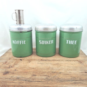 Op de afbeelding: Een set van drie groene metalen voorraadpotten met zilveren deksels, met de labels "KOFFIE", "SUIKER" en "THEE" in witte letters. De linkerpot heeft een zilveren koffiefilter. De potten staan op een houten oppervlak.