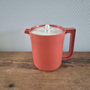 Op de afbeelding: Een rode plastic kan met een wit deksel en een witte knop bovenop. De kan heeft een handvat aan de zijkant.