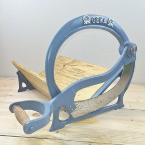 Puede incluir: Un cortador de pan de metal azul con una base de madera y un mango. El cortador tiene una cuchilla curva y las palabras "GEKA" están impresas en la parte superior.