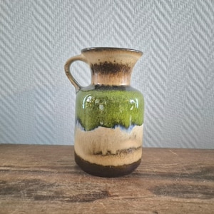 Op de afbeelding: Een keramische kan met een handvat, in bruin, beige, groen en blauw. De kan heeft een brede basis en een smalle hals, met een glanzende afwerking. Het ontwerp doet denken aan vintage aardewerk.