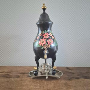 Op de afbeelding: Een zwarte metalen theepot met een bloemenmotief en een gouden tuit. De theepot heeft drie poten en staat op een zilveren standaard.