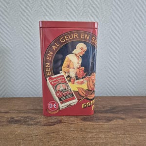 Op de afbeelding: Een rechthoekige rode metalen doos met een vintage ontwerp. De doos toont een illustratie van een vrouw die uit een theepot schenkt, met tekst in een cirkelvormige banner. Een kleinere doos met tekst en een logo is ook zichtbaar.