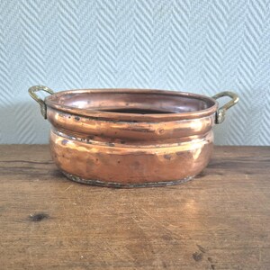 May include: A vintage copper bowl with brass handles. The bowl is round with a flared rim and a rich, reddish-brown patina. The brass handles are curved and attached to the sides. The bowl sits on a wooden surface, showing signs of age and use.