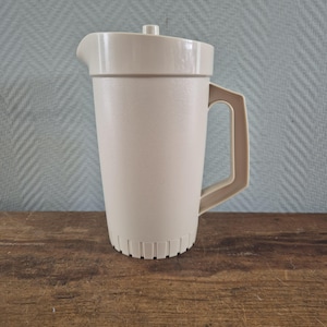 Op de afbeelding: Een beige kunststof kan met een taps toelopend cilindrisch lichaam, een stevig handvat aan de zijkant en een passend deksel met een kleine centrale knop. De onderkant van de kan is versierd met verticale ribbels en de rand heeft een schenktuit.