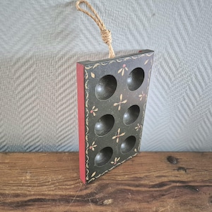 Op de afbeelding: Een houten eierrek met een zwarte achtergrond en rode rand. Het heeft een touwhandvat en is beschilderd met witte en gele bloemenmotieven. Het rek heeft 8 vakken voor eieren.