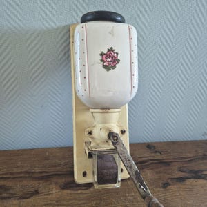 Op de afbeelding: Antieke wandkoffiemolen. De molen heeft een witte keramische container met een roze rozenontwerp en rode stippen. De basis en de hendel zijn crèmekleurig, met een glazen kijkvenster.